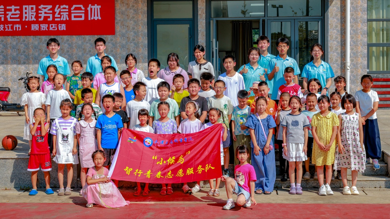 活动大合影,地点在枝江市顾家店镇养老服务综合体门前。大学生志愿者与参与暑期托管的孩子们集体合影,队伍中间展示红色旗帜:“小候鸟” 智行青春志愿服务队。标志本次乡村暑期爱心托管、七彩科技课堂活动顺利开展。