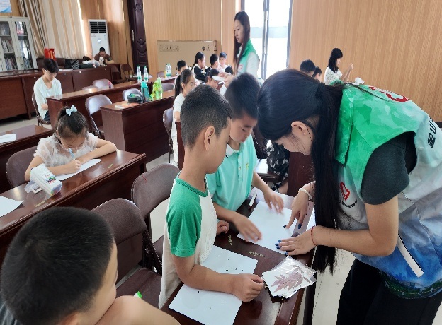 图为圆梦志愿支教队员俯身指导少年利用树叶标本开展创意手工创作。